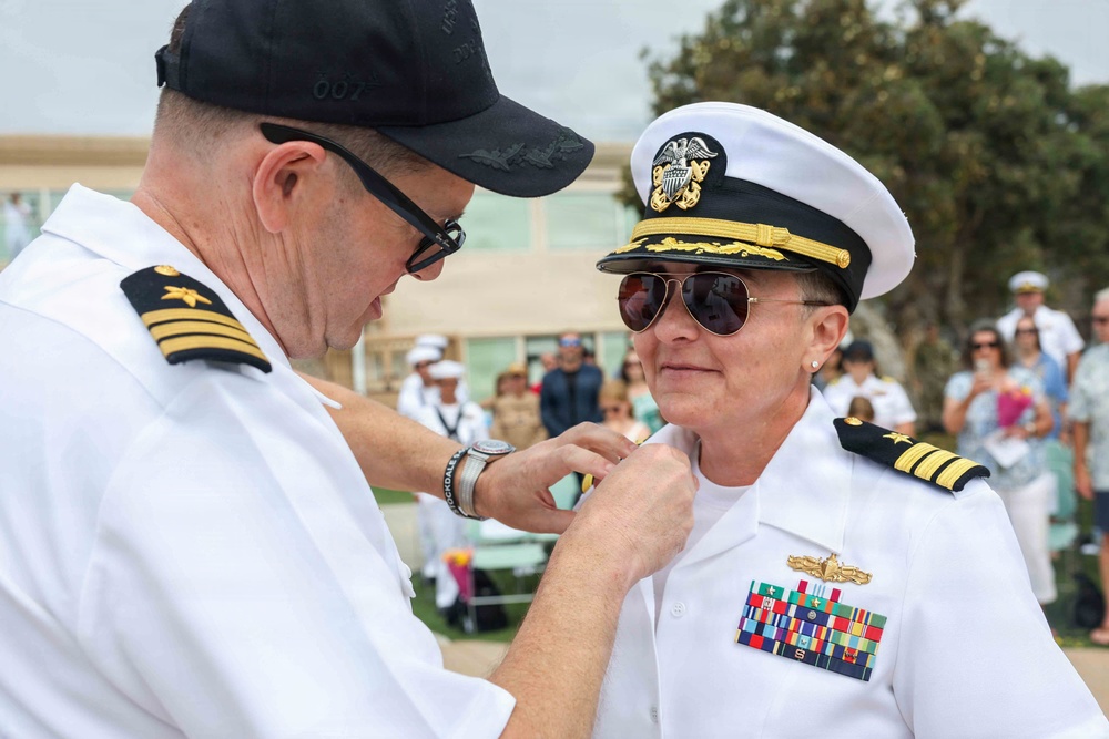 A U.S. Navy promotion or award ceremony is taking place outdoors, with one naval officer pinning a device or insignia onto another officer's uniform collar. The recipient is a female officer wearing a white dress uniform with a captain's cover, aviator sunglasses, and multiple service ribbons and a warfare qualification badge. A crowd of military personnel and civilians observes the ceremony in the background.