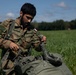 A soldier from the Qatar Armed Forces is kneeling in a green field, handling what appears to be parachute equipment or rigging lines. He is wearing camouflage uniform with visible unit patches, a name tape, and a Qatar flag patch on his sleeve. The setting appears to be a post-jump landing zone during a military airborne exercise.