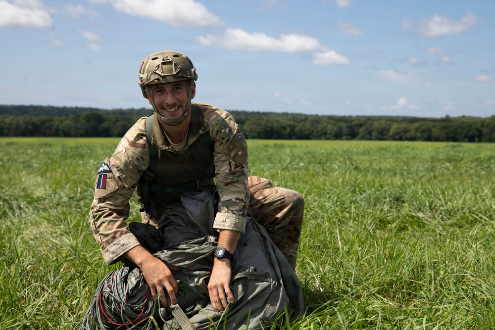A smiling soldier in MultiCam/MTP camouflage uniform is kneeling in an open green field after what appears to be a parachute landing. He is gathering parachute equipment including lines and canopy fabric. The setting is a sunny day with a tree line visible in the background.