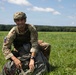 A smiling soldier in MultiCam/MTP camouflage uniform is kneeling in an open green field after what appears to be a parachute landing. He is gathering parachute equipment including lines and canopy fabric. The setting is a sunny day with a tree line visible in the background.
