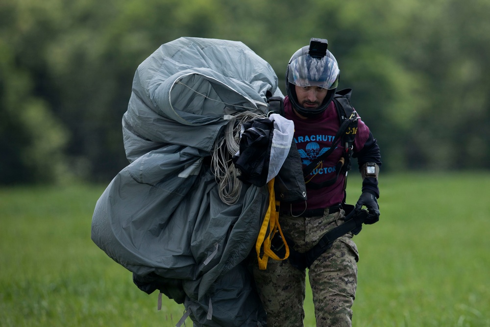 A person wearing military camouflage pants, a maroon 'Parachute Instructor' t-shirt, and a full-face helmet with a mounted camera is carrying a large bundled parachute across a grassy field. The individual appears to have just landed and is gathering and transporting the deployed parachute equipment. The setting is an open landing zone with green trees visible in the background.