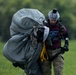 A person wearing military camouflage pants, a maroon 'Parachute Instructor' t-shirt, and a full-face helmet with a mounted camera is carrying a large bundled parachute across a grassy field. The individual appears to have just landed and is gathering and transporting the deployed parachute equipment. The setting is an open landing zone with green trees visible in the background.