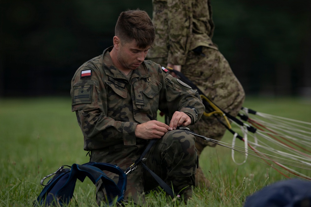 A Polish military soldier is seated on grass, carefully handling and inspecting parachute suspension lines and rigging equipment. He is wearing Polish Armed Forces camouflage (wz. 2010 pattern) with a visible Polish flag patch on his left shoulder. A second soldier in similar uniform is partially visible in the background.