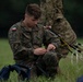 A Polish military soldier is seated on grass, carefully handling and inspecting parachute suspension lines and rigging equipment. He is wearing Polish Armed Forces camouflage (wz. 2010 pattern) with a visible Polish flag patch on his left shoulder. A second soldier in similar uniform is partially visible in the background.