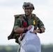 A smiling military parachutist is shown after a landing, holding a bundled white parachute canopy with mesh material and suspension lines. He is wearing full military jump gear including a tactical helmet with a mounted action camera, a parachute harness, and camouflage uniform. The background shows an open field with treeline and overcast sky.