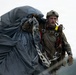 A military paratrooper is shown after a jump, carrying a large bundled parachute packed in a gray deployment bag. The soldier is wearing full tactical gear including a combat helmet, harness system, and gloves. The background suggests an overcast outdoor environment, likely a landing zone.
