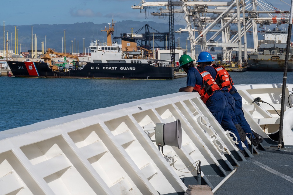 Two U.S. Coast Guard personnel in blue uniforms, orange life vests, and hard hats (green and blue) stand on the deck of a vessel, looking toward a harbor. A U.S. Coast Guard cutter bearing hull number 214 is visible in the background, moored at a port facility with large container cranes. The setting appears to be a busy commercial/military port, likely on the U.S. West Coast or Hawaii based on the terrain.