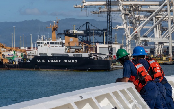 Two U.S. Coast Guard personnel in blue uniforms, orange life vests, and hard hats (green and blue) stand on the deck of a vessel, looking toward a harbor. A U.S. Coast Guard cutter bearing hull number 214 is visible in the background, moored at a port facility with large container cranes. The setting appears to be a busy commercial/military port, likely on the U.S. West Coast or Hawaii based on the terrain.