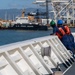 Two U.S. Coast Guard personnel in blue uniforms, orange life vests, and hard hats (green and blue) stand on the deck of a vessel, looking toward a harbor. A U.S. Coast Guard cutter bearing hull number 214 is visible in the background, moored at a port facility with large container cranes. The setting appears to be a busy commercial/military port, likely on the U.S. West Coast or Hawaii based on the terrain.