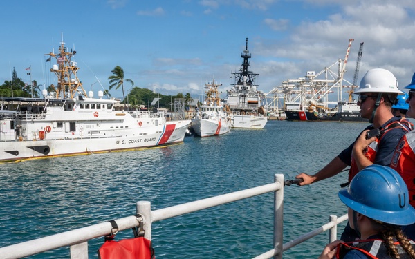 Three U.S. Coast Guard personnel wearing orange life vests and hard hats stand on the deck of a vessel, observing a harbor with multiple Coast Guard ships docked nearby. The scene appears to be at a Coast Guard station, likely in Hawaii based on the tropical vegetation and architectural style visible in the background. One crew member is holding a radio while the group monitors vessel movements in the harbor.