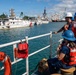 Three U.S. Coast Guard personnel wearing orange life vests and hard hats stand on the deck of a vessel, observing a harbor with multiple Coast Guard ships docked nearby. The scene appears to be at a Coast Guard station, likely in Hawaii based on the tropical vegetation and architectural style visible in the background. One crew member is holding a radio while the group monitors vessel movements in the harbor.
