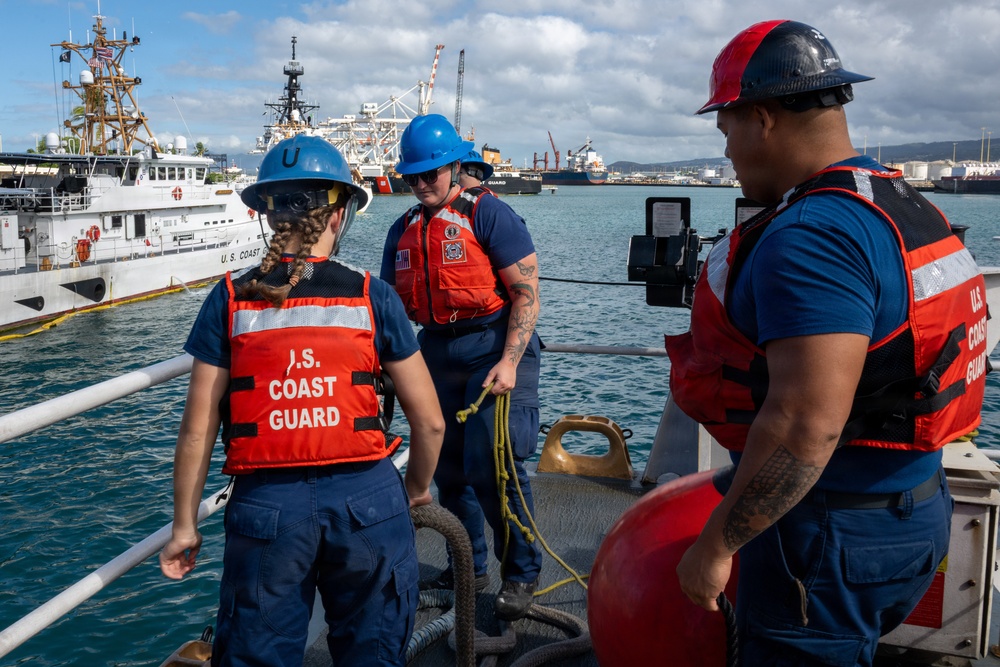Three U.S. Coast Guard personnel are aboard a vessel in a harbor, wearing orange life vests and blue or red hard hats. They appear to be conducting dockside operations, with one member handling rope/line equipment and another holding a tablet device. A white U.S. Coast Guard cutter is visible docked in the background alongside other large vessels.