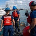 Three U.S. Coast Guard personnel are aboard a vessel in a harbor, wearing orange life vests and blue or red hard hats. They appear to be conducting dockside operations, with one member handling rope/line equipment and another holding a tablet device. A white U.S. Coast Guard cutter is visible docked in the background alongside other large vessels.