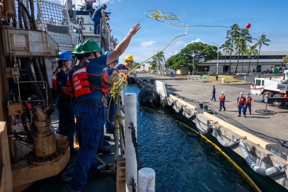 U.S. Coast Guard personnel are conducting a mooring or line-handling operation at a dock, likely at a tropical naval installation. A crew member is throwing a heaving line with a monkey fist (red ball) toward the pier while colleagues in safety gear observe. The scene depicts standard maritime berthing procedures with personnel both aboard the vessel and on the pier.