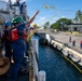 U.S. Coast Guard personnel are conducting a mooring or line-handling operation at a dock, likely at a tropical naval installation. A crew member is throwing a heaving line with a monkey fist (red ball) toward the pier while colleagues in safety gear observe. The scene depicts standard maritime berthing procedures with personnel both aboard the vessel and on the pier.