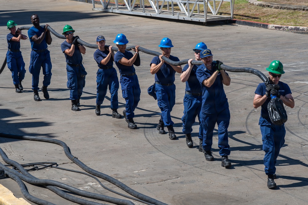 A team of approximately nine personnel wearing matching blue uniforms and hard hats are collectively carrying a large, heavy hose across an outdoor concrete surface. The group appears to be engaged in a training exercise or operational task, moving in a coordinated line formation. Their uniforms and insignia suggest they are U.S. Coast Guard members.