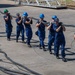 A team of approximately nine personnel wearing matching blue uniforms and hard hats are collectively carrying a large, heavy hose across an outdoor concrete surface. The group appears to be engaged in a training exercise or operational task, moving in a coordinated line formation. Their uniforms and insignia suggest they are U.S. Coast Guard members.