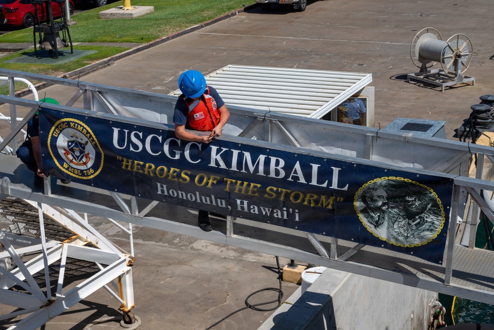 A person wearing a blue hard hat and orange life vest is securing or hanging a large navy blue banner on a railing at what appears to be a Coast Guard facility. The banner reads 'USCGC KIMBALL - Heroes of the Storm - Honolulu, Hawai'i' and displays the ship's crest (WMSL 756) along with a portrait medallion. The setting appears to be a pier or gangway area with a parking lot and green grass visible in the background.
