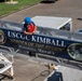 A person wearing a blue hard hat and orange life vest is securing or hanging a large navy blue banner on a railing at what appears to be a Coast Guard facility. The banner reads 'USCGC KIMBALL - Heroes of the Storm - Honolulu, Hawai'i' and displays the ship's crest (WMSL 756) along with a portrait medallion. The setting appears to be a pier or gangway area with a parking lot and green grass visible in the background.