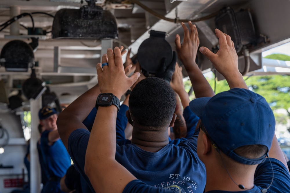 A group of personnel dressed in navy blue uniforms are working together to manipulate or install equipment mounted on an overhead structure, likely aboard a naval vessel. Multiple hands are raised upward toward what appears to be a circular hatch or mechanical component on the ceiling. The setting appears to be an interior deck or covered area of a military ship, with an instructor or supervisor visible in the background.