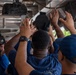 A group of personnel dressed in navy blue uniforms are working together to manipulate or install equipment mounted on an overhead structure, likely aboard a naval vessel. Multiple hands are raised upward toward what appears to be a circular hatch or mechanical component on the ceiling. The setting appears to be an interior deck or covered area of a military ship, with an instructor or supervisor visible in the background.