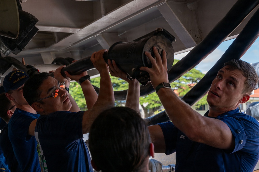 Several U.S. Coast Guard personnel in blue uniforms are working together to install or remove a large mechanical component overhead, likely aboard a vessel or in a maintenance facility. The team is engaged in a hands-on technical task requiring coordinated effort. At least one person's cap is visible with 'USCGC KIMBALL' text, identifying the unit.