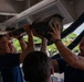 Several U.S. Coast Guard personnel in blue uniforms are working together to install or remove a large mechanical component overhead, likely aboard a vessel or in a maintenance facility. The team is engaged in a hands-on technical task requiring coordinated effort. At least one person's cap is visible with 'USCGC KIMBALL' text, identifying the unit.