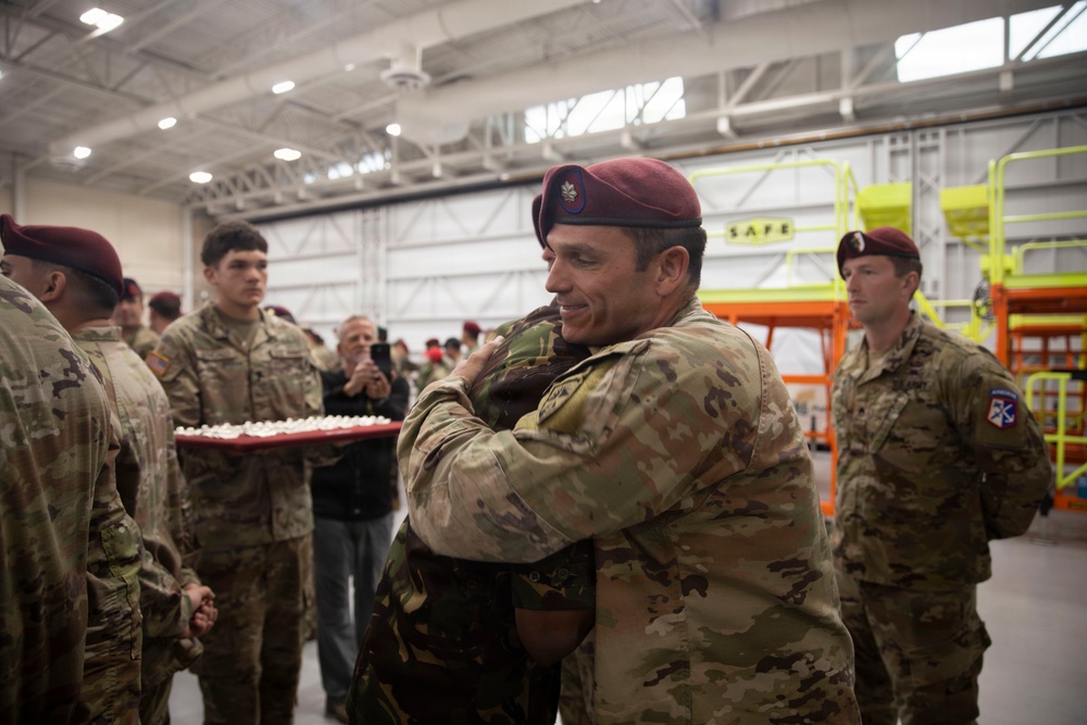 A military ceremony taking place inside what appears to be an aircraft hangar, where two service members are embracing in what looks like a farewell or congratulatory moment. Multiple soldiers wearing maroon berets and MultiCam/OCP uniforms are present, suggesting a unit associated with airborne or special operations forces. A red tray with what appears to be cupcakes or small items is visible in the background, indicating a celebratory occasion.