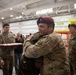 A military ceremony taking place inside what appears to be an aircraft hangar, where two service members are embracing in what looks like a farewell or congratulatory moment. Multiple soldiers wearing maroon berets and MultiCam/OCP uniforms are present, suggesting a unit associated with airborne or special operations forces. A red tray with what appears to be cupcakes or small items is visible in the background, indicating a celebratory occasion.