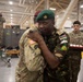 A Kenyan military officer wearing a green beret and Kenya flag patch embraces a U.S. Army soldier in a military facility or hangar. The scene appears to be a multinational military exchange or ceremony, evidenced by the presence of multiple soldiers and a tray of what appear to be military coins or medals. A Union Jack (British flag) is visible hanging in the background, suggesting a joint allied military event.