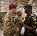 Two military officers from different nations are shaking hands in what appears to be a large indoor military facility or hangar. The officer on the left wears a US Army uniform with a maroon beret and Ranger tab, while the officer on the right wears a Kenyan military uniform with a green beret and Kenya flag patch. Several other military personnel are visible in the background, along with national flags including the United Kingdom and Spain.