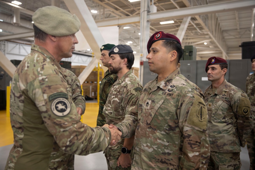 A military ceremony or greeting is taking place inside what appears to be a large hangar or military facility. A senior officer wearing a green beret is shaking hands with a U.S. Army soldier wearing a maroon beret, while other military personnel in mixed uniforms stand in formation nearby. The personnel appear to represent multiple military units or possibly allied nations.