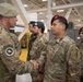 A military ceremony or greeting is taking place inside what appears to be a large hangar or military facility. A senior officer wearing a green beret is shaking hands with a U.S. Army soldier wearing a maroon beret, while other military personnel in mixed uniforms stand in formation nearby. The personnel appear to represent multiple military units or possibly allied nations.