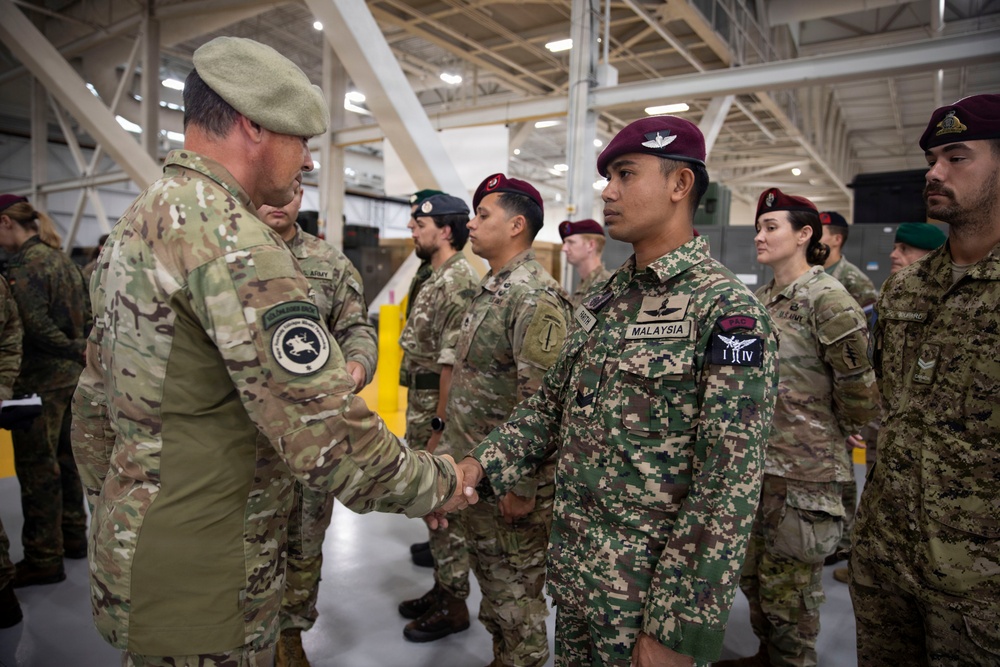 A military ceremony or greeting event taking place inside a large hangar or military facility. A senior officer wearing a green beret is shaking hands with a Malaysian soldier in digital camouflage uniform, while a line of multinational military personnel stands at attention in the background. The scene appears to be a multinational military exercise or joint training event.