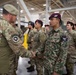 A military ceremony or greeting event taking place inside a large hangar or military facility. A senior officer wearing a green beret is shaking hands with a Malaysian soldier in digital camouflage uniform, while a line of multinational military personnel stands at attention in the background. The scene appears to be a multinational military exercise or joint training event.