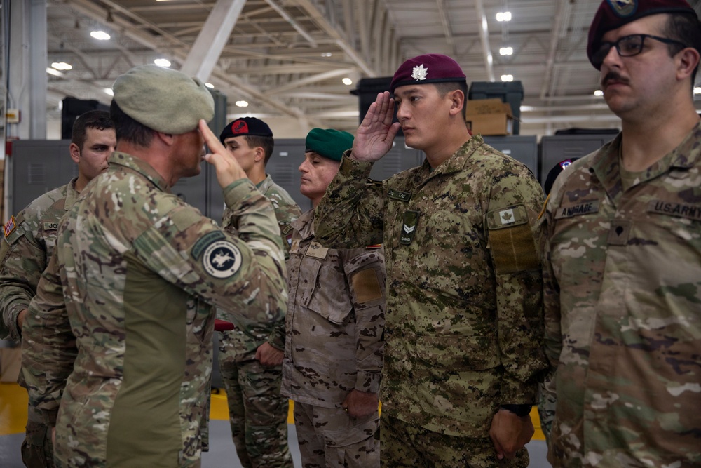 A multinational military ceremony is taking place inside what appears to be a large indoor facility or hangar. Soldiers from multiple nations, identifiable by different uniforms and beret colors, are exchanging salutes. The scene suggests a formal military event such as an award ceremony, inspection, or unit exchange.