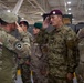 A multinational military ceremony is taking place inside what appears to be a large indoor facility or hangar. Soldiers from multiple nations, identifiable by different uniforms and beret colors, are exchanging salutes. The scene suggests a formal military event such as an award ceremony, inspection, or unit exchange.