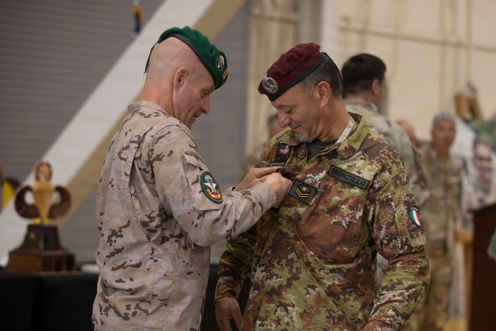 Two military personnel are engaged in a medal or decoration pinning ceremony. The officer on the left, wearing a green beret and desert camouflage uniform, is pinning something onto the chest of the officer on the right, who wears a maroon beret and Italian digital camouflage uniform with a name tape reading 'MONZILLO' and an Italian flag patch. The setting appears to be an indoor military facility, likely a hangar, with trophies and other personnel visible in the background.