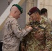 Two military personnel are engaged in a medal or decoration pinning ceremony. The officer on the left, wearing a green beret and desert camouflage uniform, is pinning something onto the chest of the officer on the right, who wears a maroon beret and Italian digital camouflage uniform with a name tape reading 'MONZILLO' and an Italian flag patch. The setting appears to be an indoor military facility, likely a hangar, with trophies and other personnel visible in the background.
