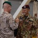Two military personnel from different nations are shaking hands and smiling in what appears to be a military facility or hangar. The soldier on the right is visibly identified as Italian Army (Esercito) with the name tag 'Monzillo', wearing a maroon paratrooper beret, while the soldier on the left wears a green beret and desert camouflage uniform. The interaction appears cordial and celebratory, suggesting a multinational military event or ceremony.