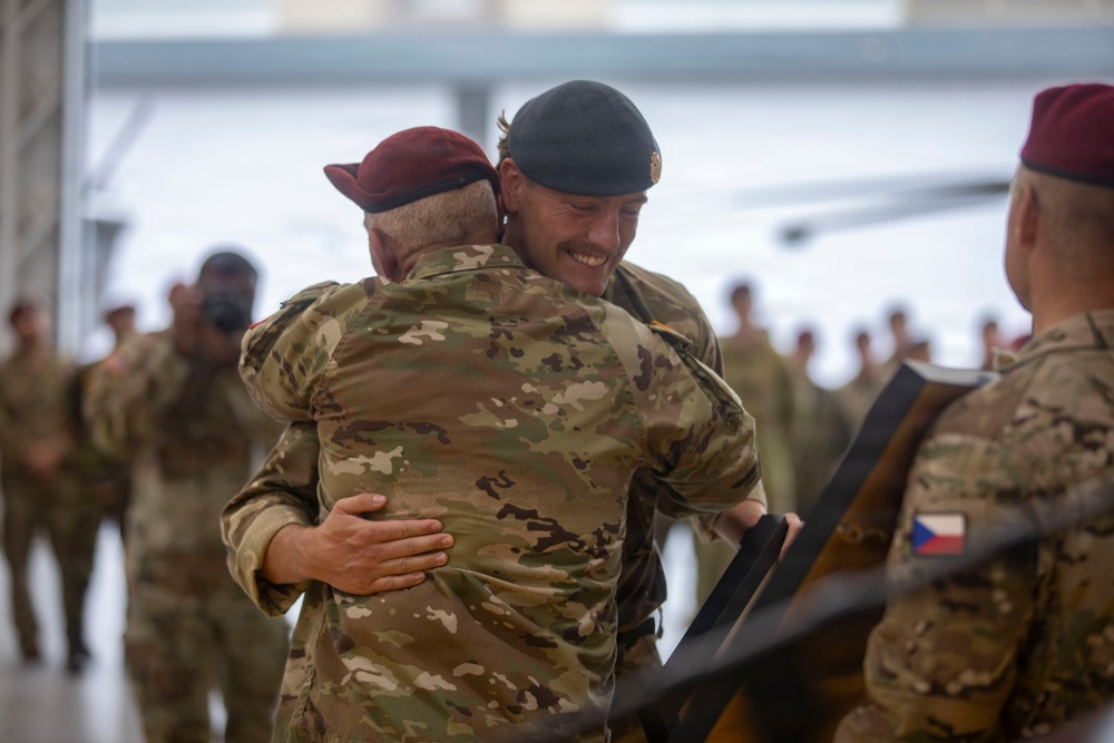 Two military personnel in camouflage uniforms are embracing in what appears to be a hangar or large military facility. One soldier wears a dark/black beret and is smiling broadly, while the other wears a maroon/burgundy beret. A third soldier visible on the right displays a Czech Republic flag patch on their uniform.