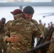 Two military personnel in camouflage uniforms are embracing in what appears to be a hangar or large military facility. One soldier wears a dark/black beret and is smiling broadly, while the other wears a maroon/burgundy beret. A third soldier visible on the right displays a Czech Republic flag patch on their uniform.