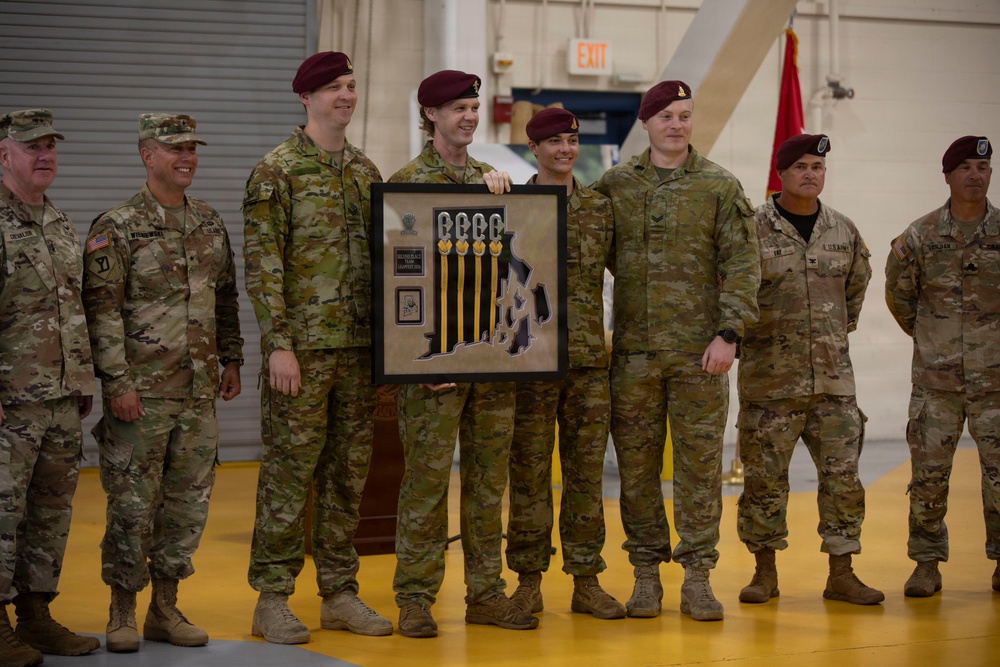 A group of military personnel in U.S. Army camouflage uniforms pose together during what appears to be an awards or recognition ceremony held inside a military hangar or gymnasium. Several individuals are wearing maroon berets, indicative of airborne/paratrooper units, while others wear patrol caps. Two central figures are holding a framed commemorative plaque or shadow box with medals and insignia.