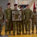 A group of military personnel in U.S. Army camouflage uniforms pose together during what appears to be an awards or recognition ceremony held inside a military hangar or gymnasium. Several individuals are wearing maroon berets, indicative of airborne/paratrooper units, while others wear patrol caps. Two central figures are holding a framed commemorative plaque or shadow box with medals and insignia.