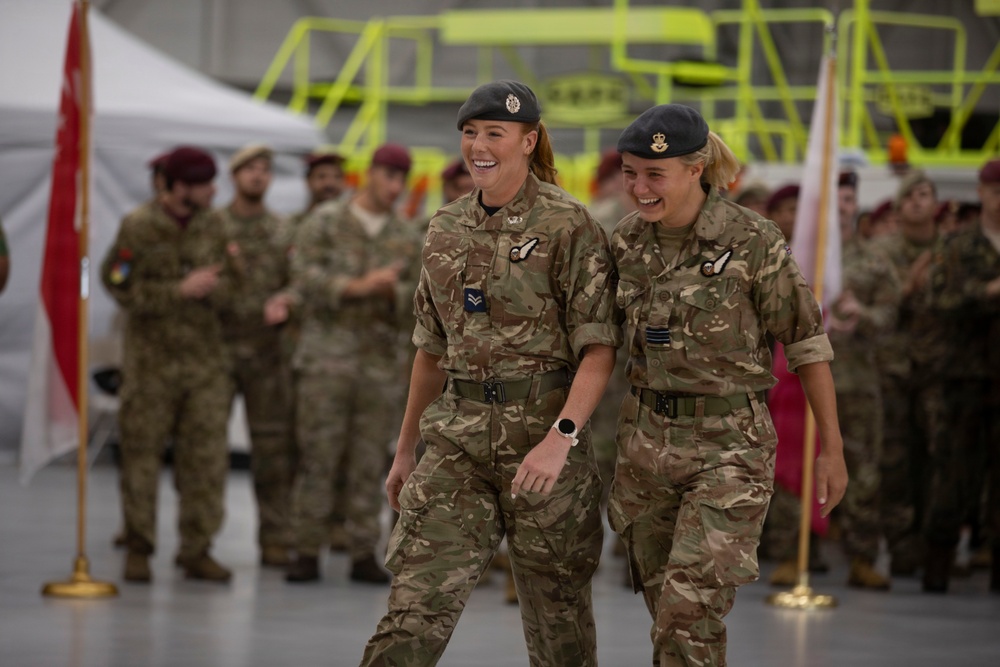Two female military personnel in British Army MultiTerrain Pattern (MTP) camouflage uniforms are walking and laughing together during what appears to be a formal military ceremony or parade held inside an aircraft hangar. Both wear black berets with cap badges and have wing-style qualification badges on their uniforms. A group of mixed military personnel, including some wearing red berets, are visible in the background.