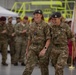 Two female military personnel in British Army MultiTerrain Pattern (MTP) camouflage uniforms are walking and laughing together during what appears to be a formal military ceremony or parade held inside an aircraft hangar. Both wear black berets with cap badges and have wing-style qualification badges on their uniforms. A group of mixed military personnel, including some wearing red berets, are visible in the background.