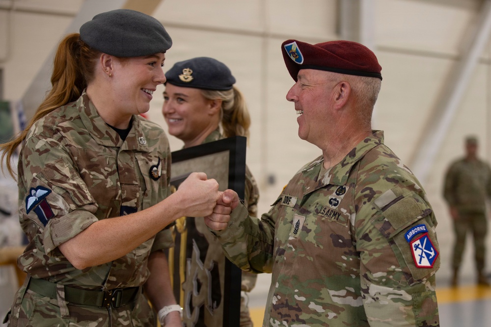 A British Army soldier and a U.S. Army soldier are sharing a fist bump and smiling in what appears to be a military hangar or indoor facility. A second British soldier is visible in the background, also smiling. The interaction suggests a friendly, ceremonial, or congratulatory exchange between allied military personnel.