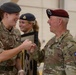 A British Army soldier and a U.S. Army soldier are sharing a fist bump and smiling in what appears to be a military hangar or indoor facility. A second British soldier is visible in the background, also smiling. The interaction suggests a friendly, ceremonial, or congratulatory exchange between allied military personnel.