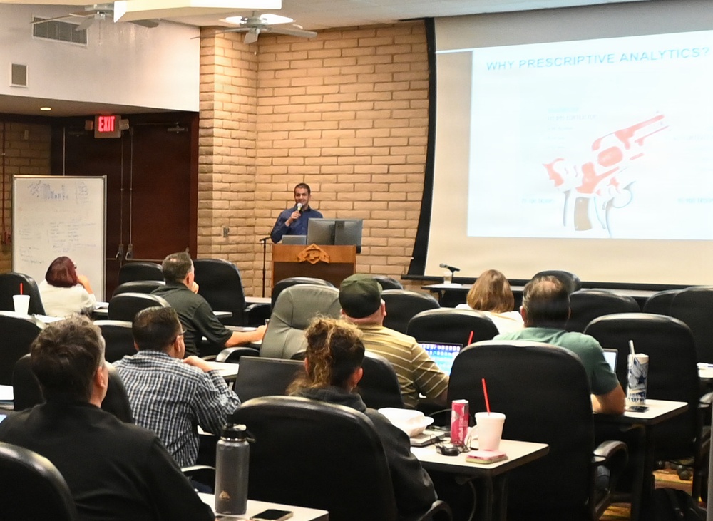 A presenter is delivering a lecture or seminar in a tiered classroom or auditorium setting. The projected slide displays the title 'WHY PRESCRIPTIVE ANALYTICS?' with accompanying visuals. An audience of approximately 10-15 attendees is seated at individual desks, some with laptops and beverages.