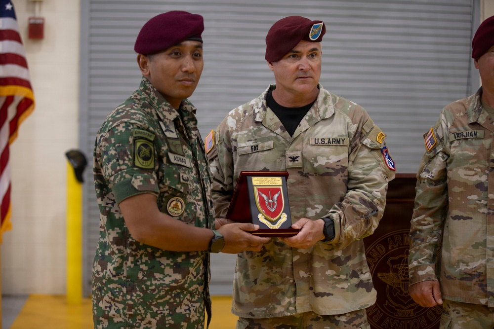 Two military personnel are jointly holding a commemorative plaque during what appears to be a formal ceremony. The soldier on the left wears a foreign military uniform with the name tag 'ASWAD' and an airborne patch, while the U.S. Army soldier on the right bears the name tag 'FAY' and wears the rank of Colonel (eagle insignia). A third soldier partially visible on the right has the name tag 'VARJIAN'.